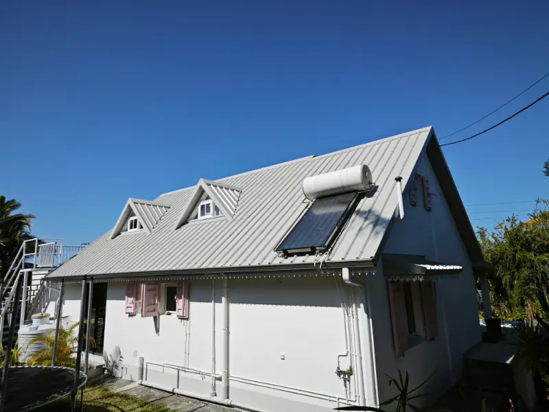 Toiture en bac acier avec deux lucarnes et un chauffe-eau solaire, sous un ciel bleu.