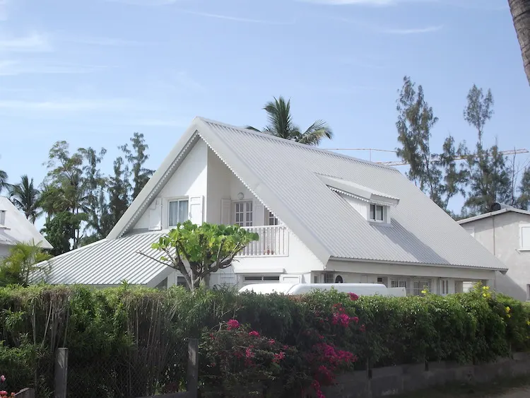 Maison à étage au toit de tôle blanche et à la lucarne, derrière une haie fleurie.